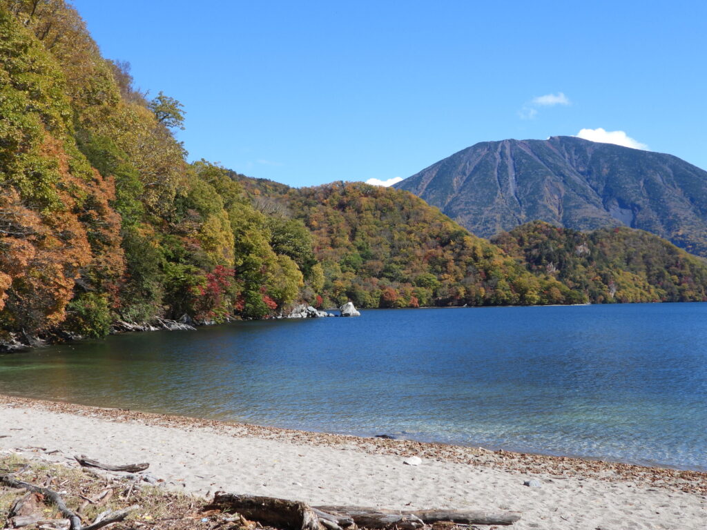 Autumn scenery of Lake Chuzenji in Nikko National Park, with clear blue water, forested mountains, and colorful fall foliage along the shoreline.